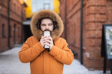 Kışın parka giyen adam termosu kapalı bir şekilde karlı şehir sokağında tutuyor. Soğuk havada sıcaklık, yalnızlık ve konfor.