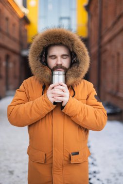 Kürklü adam, karlı caddede sıcak içecekten hoşlanıyor. Huzurlu yalnızlık, kış rutini ve soğuk havada şehir hayatı..