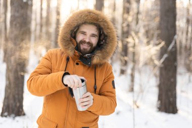 Adam karlı kış ormanlarında termosla ısınıyor. Soğuk doğal ortamda konfor ve sabah farkındalığı.