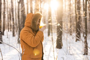 Karlı ormanda sarı bardaktan içen adam. Kışın hayatta kalmasını, günlük ritüelleri ve dışarıdaki sıcaklıkla farkındalık arasındaki bağlantıyı yansıtır.