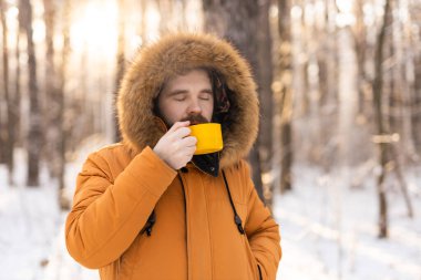 Karlı ormanda sarı bardaktan içen adam. Kışın hayatta kalmasını, günlük ritüelleri ve dışarıdaki sıcaklıkla farkındalık arasındaki bağlantıyı yansıtır.