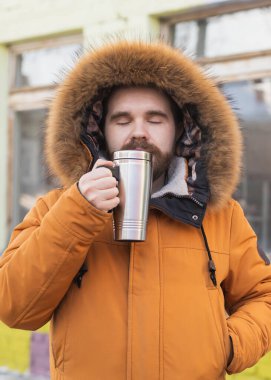 Close-up portrait handsome bearded millennial man in winter clothes and with thermos snow outdoor. Cold season and hot beverage in winter time.