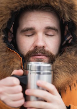 Close-up portrait handsome bearded millennial man in winter clothes and with thermos snow outdoor. Cold season and hot beverage in winter time.