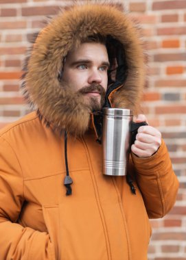 Kış sokağında termoslu bir adam tuğla duvarın yanında duruyor. Kentsel yaşam tarzı, mevsimsel rutin ve soğuk havada açık havada rahatlama.