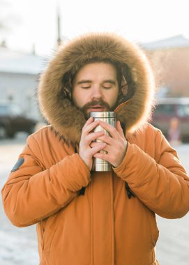 Close-up portrait handsome bearded millennial man in winter clothes and with thermos snow outdoor. Cold season and hot beverage in winter time.