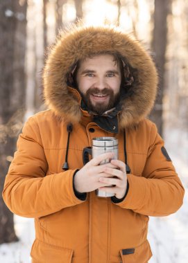 Close-up portrait handsome bearded millennial man in winter clothes and with thermos snow outdoor. Cold season and hot beverage in winter time.