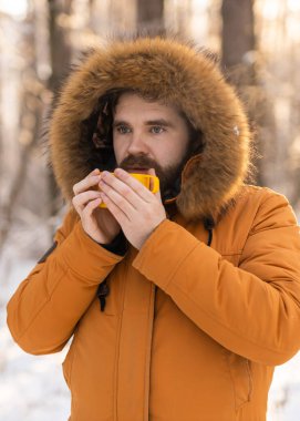 Karlı ormanda sarı bardaktan içen adam. Kışın hayatta kalmasını, günlük ritüelleri ve dışarıdaki sıcaklıkla farkındalık arasındaki bağlantıyı yansıtır.