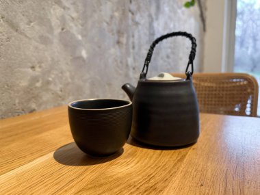 Sıcak kafenin iç tarafındaki ahşap masada fincanlı siyah seramik çaydanlık. Sakin, ritüel ve dikkatli çay içme deneyimi..