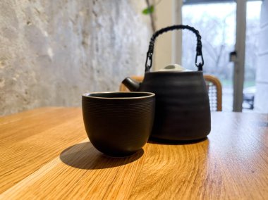 Sıcak kafenin iç tarafındaki ahşap masada fincanlı siyah seramik çaydanlık. Sakin, ritüel ve dikkatli çay içme deneyimi..