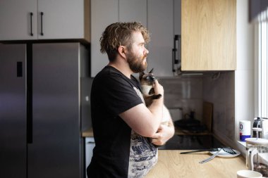 Bir adam kediyi sessiz bir mutfakta pencere ışığında tutuyor. Rahatlık, evcil hayvan duygusal bağ ve günlük ev hayatının huzurlu ritmi..