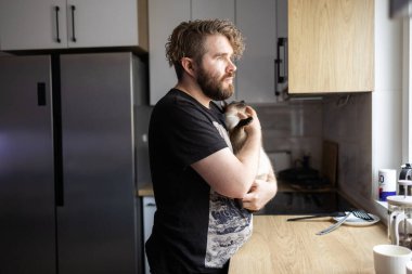 Bir adam kediyi sessiz bir mutfakta pencere ışığında tutuyor. Rahatlık, evcil hayvan duygusal bağ ve günlük ev hayatının huzurlu ritmi..