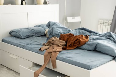 Yatak odası sahnesinde dağınık giysilerle yapılmamış bir yatak. Ev işleri, günlük ihmaller ve ev hayatının sessiz kaosu..