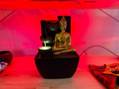 Kırmızı ışığın altında masa başı çeşmesi olan altın Buda heykeli. İç huzur, ruhani uygulama ve kutsal bir mekanda meditasyon varlığı..
