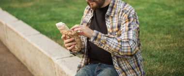 Adam açık havada kağıdın içinde sokak yemeği tutuyor. Fast food kavramı, günlük yaşam tarzı ve şehir parkında atıştırma..