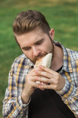 Adam öğle yemeği molasında sandviç paketini ısırıyor. Günlük fast food kavramı, sokak yemekleri kültürü ve açık havada basit zevklerin tadını çıkarmak.