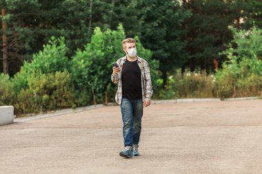 Parkta yürürken maske takan ve akıllı telefonunu kontrol eden bir adam. Modern yaşam, sağlık güvenliği ve salgın zamanlarında iletişim kavramı. Boşluğu kopyala.