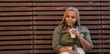 Afrika kökenli Amerikalı bir kadın kentteki bir bankta otururken pipetle içkisini yudumluyor. Dinlenme, rahatlama ve basit günlük anlar..