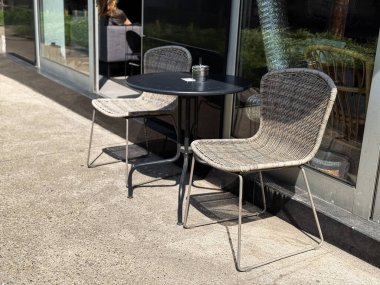 İki hasır sandalyeli açık kafe masası. Kaldırımda küçük yuvarlak bir masa, parlak güneş ışığı altında cam kül tablası.