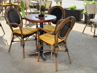 Sarı yastıklı açık kafe koltuklar. Yuvarlak ahşap masa, rattan sandalyeler ve saksı bitkisi süsü.