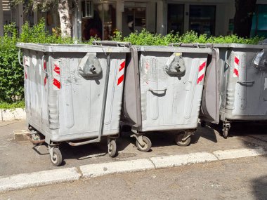 Şehir caddesinde bir sıra metal çöp tenekesi. Kaldırımların ve yeşilliklerin yanına tekerlekleri olan endüstriyel atık konteynırları