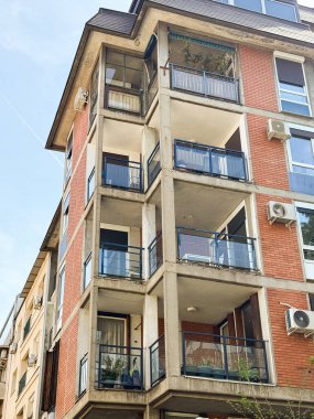 Balkonlu ve kırmızı tuğlalı bir bina. Cam korkuluklar ve havalandırma ünitelerinin yer aldığı çok katlı bir apartman.