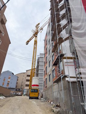 Tamamlanmamış apartmanın yanında uzun bir vinç. Binanın etrafı inşaat malzemeleri ve toprak bir yol ile çevrili.