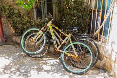 Gölgeli sokakta duvara yaslanmış iki eski model bisiklet. Kentsel bozulma, nostaljik ulaşım ve kırsal Akdeniz cazibesi.
