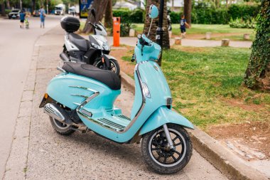 Turkuaz scooter yeşil parkın yakınındaki bir caddeye park edilmiş. Kentsel hareketlilik, kişisel ulaşım ve yaz tatili