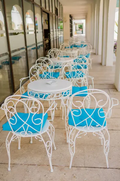 Güneşli verandada mavi yastıklı beyaz teras sandalyeleri. Açık hava zarafeti, Akdeniz eğlencesi ve yaz konukseverliği tasarımı.