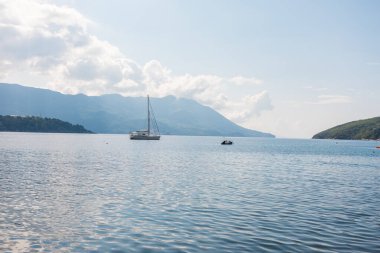 Yelkenli, yumuşak güneş ışığı altında dağlık kıyı şeridine demir attı. Sahil seyahati, deniz eğlencesi ve manzaralı yaz kaçışı.