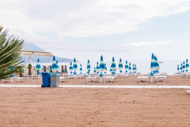 Empty beach with rows of closed umbrellas and sun loungers on a cloudy day. Coastal relaxation, off-season atmosphere and summer tourism infrastructure