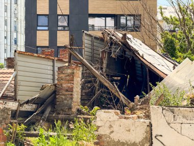 Yıkık çatılı ve etrafı yabani otlarla çevrili yıkılmış bir ev. Çürüme, yıkım ve geçmiş ve şimdiki mimarinin karşıtlığı