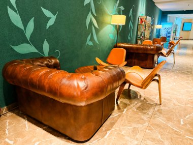 Deri koltuklar ve ahşap sandalyeler modern lobide hafif ışıklandırmayla dizilmiş. Konukseverlik, konfor ve iç tasarım rahatlamayı, yaşam tarzını ve çağdaş atmosferi sembolize eder.