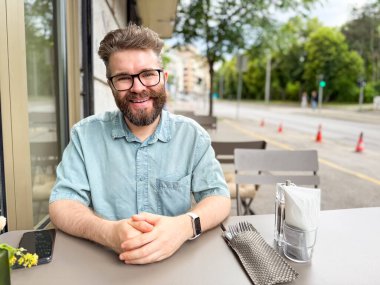 Dışarıdaki kafe masasında oturan sakallı adam yan tarafa bakıyor. Modern yaşam tarzı, şehir eğlencesi ve günlük sosyal çevrenin rahat atmosferi.