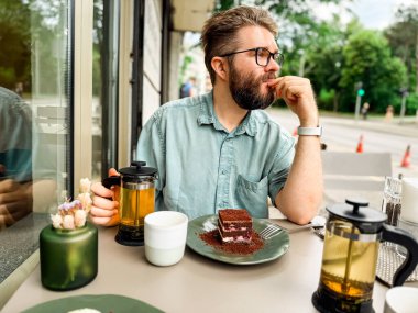 Dışarıdaki kafe masasında oturan sakallı adam yan tarafa bakıyor. Modern yaşam tarzı, şehir eğlencesi ve günlük sosyal çevrenin rahat atmosferi.