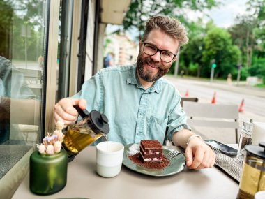 Dışarıdaki kafe masasında gülümseyen bir adam fincana çay dolduruyor. Kentsel ortamlarda sosyal yaşam tarzı rahatlama, yiyecek ve içecek keyfi