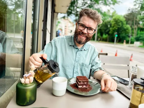 Dışarıdaki kafe masasında gülümseyen bir adam fincana çay dolduruyor. Kentsel ortamlarda sosyal yaşam tarzı rahatlama, yiyecek ve içecek keyfi