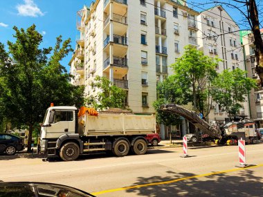 İnşaat kamyonu ve asfalt kaldırım taşıtları şehir caddesinde konut binalarının yanında çalışıyor. Kentsel yol çalışması ve ağır ekipman operasyonu altyapı bakımı ve ilerlemeyi sembolize ediyor