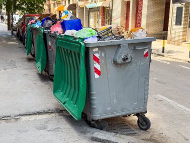 Şehir caddesinde park halindeki arabaların yanında duran çöp bidonları. Kentsel temizlik, atık yönetimi ve geri dönüşüm altyapısı kirliliği, çevreyi ve belediye hizmetlerini sembolize ediyor