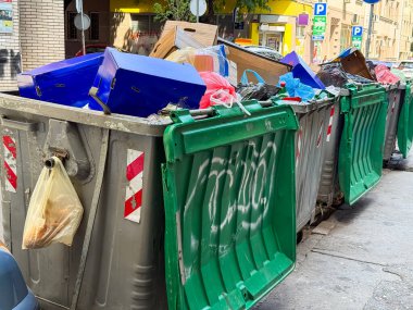 Şehir caddesinde park halindeki arabaların yanında duran çöp bidonları. Kentsel temizlik, atık yönetimi ve geri dönüşüm altyapısı kirliliği, çevreyi ve belediye hizmetlerini sembolize ediyor