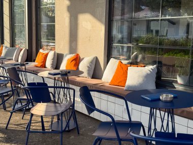 Mavi masaların yanındaki banklarda turuncu ve beyaz yastıklı modern kafe terası. Konfor, rahatlık ve sosyal etkileşim çağdaş konukseverlik ve açık hava kültürü yoluyla