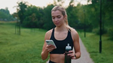 Sportif beyaz kadın dışarı çıkıyor, akıllı telefon kullanıyor spor su şişesini tutuyor, mesajlaşıyor, uygulamaları kaydırıyor. Parkta yürürken internette gezinen bayan sporcu. Sağlıklı yaşam tarzı kavramı