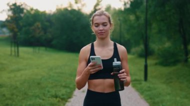 Çekici sporcu Kafkasyalı koşucu kadın parka gidiyor, akıllı telefon kullanıyor spor su şişesini tutuyor, mesajlaşıyor, yazışıyor.