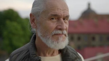 Şehrin dışında duran, gülümseyen, spor elbiseli, olgun bir adamın portresi bulanık arka planda. Yaşlı stil sahibi erkek emekli kameraya bakıyor. Genççe yaşlanıyor. Kapat.