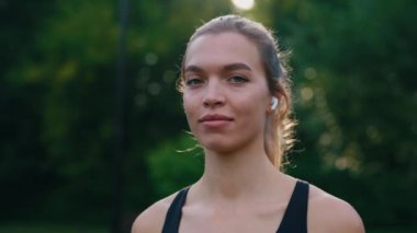 Dışarıda duran çekici bayan koşucunun portresi. Arka plandaki kameraya bakıyor. Kulaklık takıyor. Yaz zamanı. İnsanlar ve spor anlayışı. Kapat.