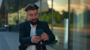 İş merkezinin yanında duran sakallı erkek yönetici ellerini elektrikli scooter 'a koyup telefonu kullanıyor. Arkadaşlarıyla iletişim kuruyor. Müşterilerle uyuşuyor. Oyun oynuyorum.