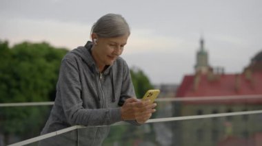 Olgun bir kadın dışarıda akıllı telefon kullanıyor, mesaj atıyor, kulaklık takıyor. Eğitimden sonra mesajlaşan yaşlı bayan sporcu. İnsanlar, spor ve teknoloji kavramı