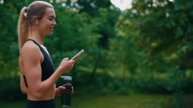 Spor kadını yürürken, spor şişesini tutarken, telefonu kaydırırken ve kulaklıkla müzik dinlerken yan görüntüsü. Spor ve teknoloji ve sağlıklı yaşam tarzı konsepti