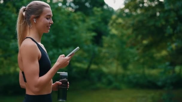 Spor Kadını Yürürken Spor Şişesini Tutarken Telefonu Kaydırırken Kulaklıkla Müzik — Stok video