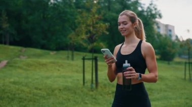 Sportif beyaz kadın parka gidiyor, akıllı telefon kullanıyor, spor su şişesini tutuyor, şık giyiniyor. Spor ve insanlar konsepti